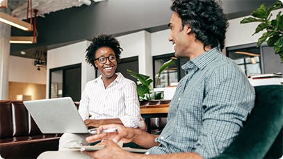 Two people sitting indoors smiling. The person in the background is using a laptop.