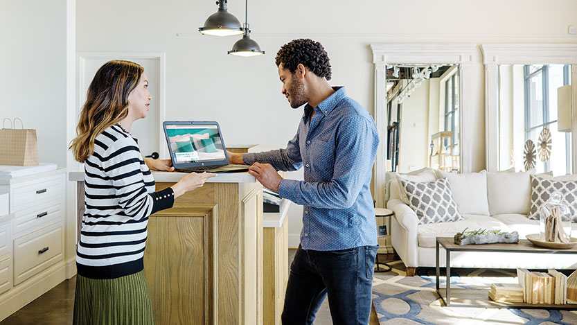 Two people stand in a home kitchen looking at a shopping website on a laptop computer.