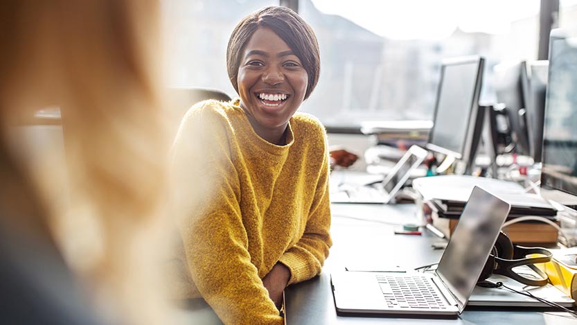 A woman wearing a yellow sweater sits at an office in front of  laptop and smiles while talking to someone else.