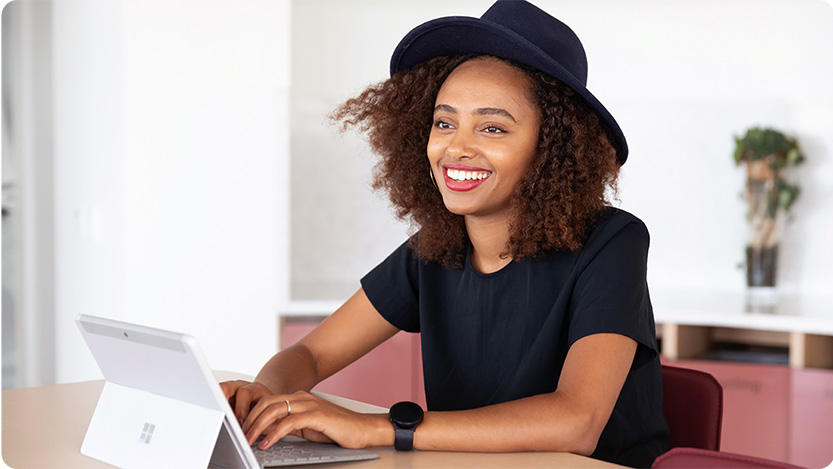 A person with a black t-shirt and a black hat, sitting at a table, smiling, and using a tablet.