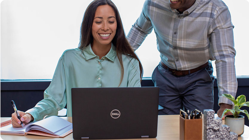 Two people, one is sitting at a desk taking notes in a notebook while looking and smiling at a laptop. The second person in standing on the side and smiling.